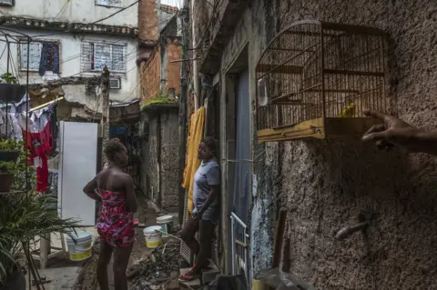 Tariq Zaidi Women waiting in one of the alleyways while they fill up their water buckets. Favela Vila do Metro community, Mangueira, Rio de Janeiro, Brazil