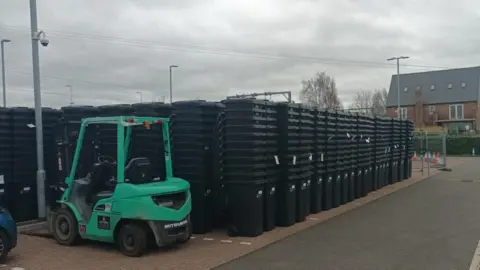 East Cambridgeshire District Council Rows of new black bins, stacked in piles of 11 are on a car park. To one side is a green forklift truck.