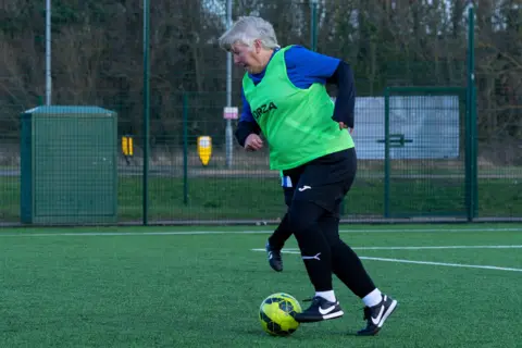 Simon Thurlow Lauri Chandler has short white hair, blue and black sports top, green bib, black sports bottoms and black football boots. She is dribbling a yellow and black football. She is on on a football pitch surrounded by a green cage.