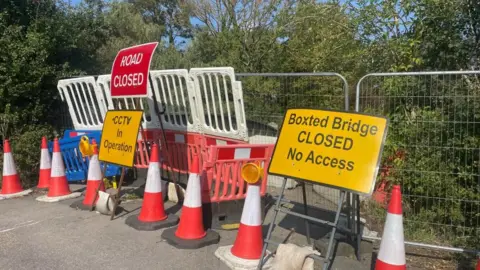 Picture of white barriers and metal fencing with red and white bollards and road signs in front. There is greenery in the background.