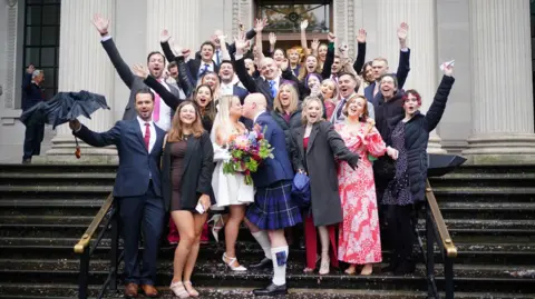 PA MEDIA People crowd and cheer on the steps of the old marlyebone town hall as thomas and paige mackintosh celebrate tying the knot on 1 October. Confetti can be seen on the floor while the group have their hands in the air cheering