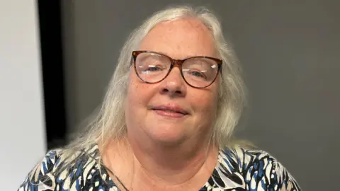 Jane Johnson, who has shoulder-length grey hair, wears brown glasses. She is photographed wearing a patterned white, dark blue and black top.