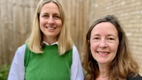 Mark Norman / BBC Two woman face the camera, Dr Rodder, on the left has long blonde hair and is wearing a green jumper and white blouse, Professor Claudia Cooper on the right of the picture has long brown hair and is wearing a black blouse. Both woman are standing outside in front of a wooden fence and a brick wall