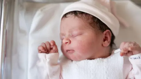 A newborn baby wearing a pink top and a pink hat sleeps in a hospital cot