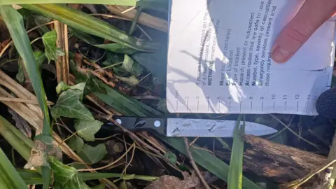West Midlands Police A knife lying in greenery, with a hand positioning a paper ruler next to the blade to measure it.