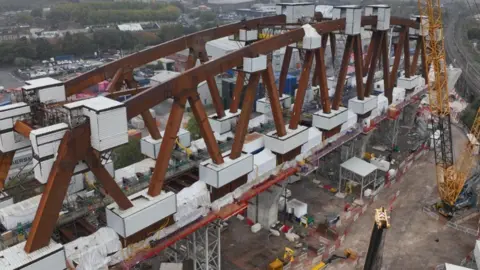 HS2 Ltd Looking down onto a huge steel truss with construction happening around it. There are white boxes on the joins. In the background you can see trees and cars. Below is dirt, fencing and equipment, including a yellow crane to the right. 