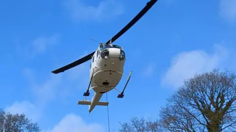 The helicopter hovering over a black cylinder that it's delivering. Workers in orange PPE are helping with the delivery