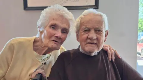 Sylvia, 88, and her 92-year-old husband Fred. She has wispy short grey hair and is wearing a yellow cardigan. She has her arm around Fred, who has short grey hair and is wearing a maroon jumper.