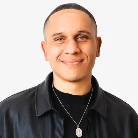 Thomas Morgan A man wearing a black leather jacket and a silver necklace smiles at the camera