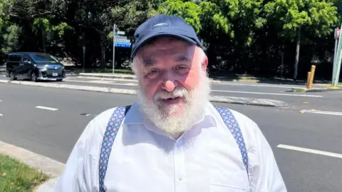 ABC News: Danuta Kozaki An elderly man with a bushy white beard and a baseball cap smiles at the camera
