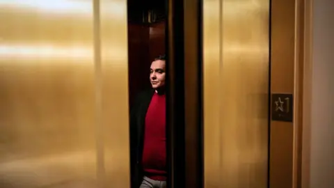 Drew Angerer George Santos in red sweater between gold elevator doors.