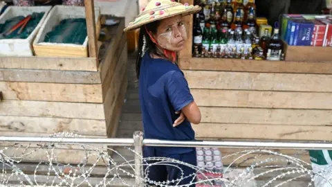 A Myanmar vendor (C) selling alcohol, cigarettes and aphrodisiac looks on while waiting for customers inside a "no-man's land" between Thailand and Myanmar, as seen from behind a barb-wire fence in Thailand's Mae Sot district on April 12, 2024
