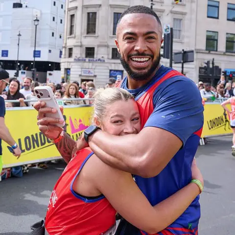 Getty Images Tyler West and Molly Rainford smile and embrace for a photo while holding a smartphone during the London Landmarks Half Marathon event.