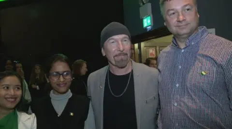 RTÉ David Howell Evans, with beard and grey hat, grey jacket and necklace, third from the left. He is accompanied on his left, by a woman with dark hair, green top and white jacket, a woman with dark hair, black top and grey jumper and a man with grey hair and red/blue striped shirt.