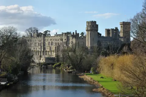 An image of Warwick's grey stone medieval castle taken from a bridge over the river Avon
