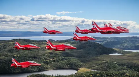 MOD Red Arrows team flying over a vast Canadian landscape, carpeted with greenery. The team is in formation.