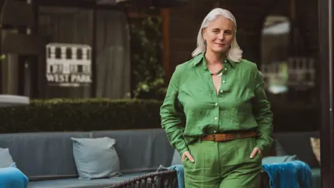 Eva Boruc Photography A woman with grey hair walks through an outdoor patio area wearing a bright green shirt and matching green trousers, with a brown belt and shoes.