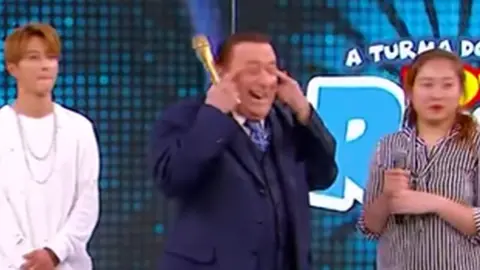 A still from YouTube showing Brazilian TV host Raul Gil making a "slant eye" gesture while interviewing Korean pop group KARD