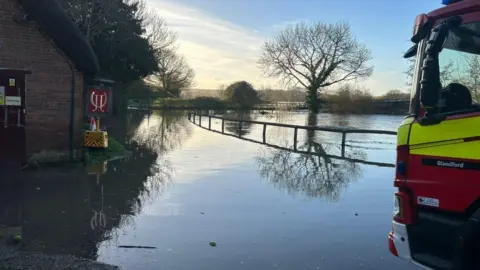 Dorset & Wiltshire Fire and Rescue Service Fire engine stopped on edge of flooded road with building on left and wooden fence on right.