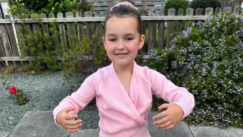 Family image shows the image of Baby King wearing a pink ballet uniform and standing in the ballet in a garden.