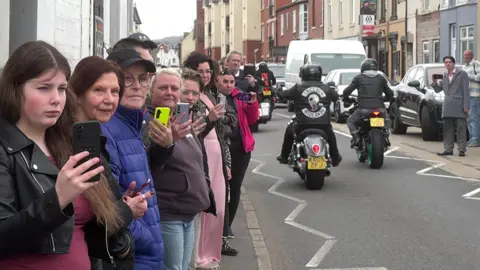 A number of people lining the street, many have phones which they are using to film the procession. There are motorbikes in the background, as if they have just driven past as part of proceedings. 