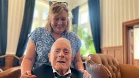 Robin Boodle is in the lower half of the frame looking at the camera, wearing a black suit and tie. His daughter, Gay Kilshaw, is standing with her hands on his shoulders looking down at him. she is wearing a blue and white dress.