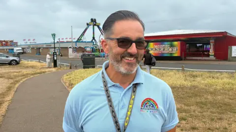 Robby West/BBC Anthony Parkin smiles towards the camera, while wearing a light blue polo top, sunglasses and an Oakley branded lanyard. He is stood in front of the entrance to a fairground