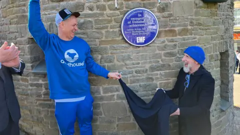 James Atkin dressed in a royal blue-coloured jumper and baseball cap and Ian Dench in a black coat and royal blue beanie, pulling the sheet from their plaque on Cinderford's clock tower. It reads 'Contribution to Music. In recognition of Unbelievable achievements, EMF'