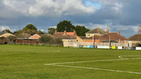 Ben Hails A grass football pitch is shown with houses in the background beyond a fence.