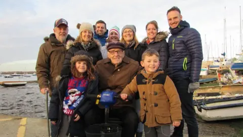 Elderly man in a wheelchair with relatives of various ages including children next to a river