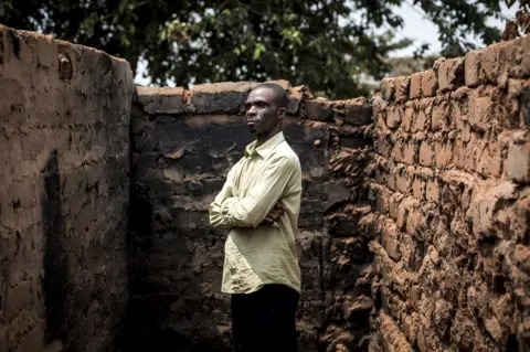 John Wessels / Oxfam Emmanuel stands in front of the remains of his house.