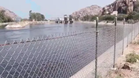 PTI Blue water flows in the Sanapur river canal near Hampi. We can see a wire railing supported by metal pipes and boulders in the background. 