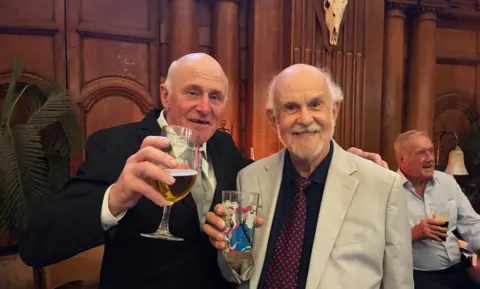 Maxine Beuret Two men stand next to each other. The man on the left has his hand on the shoulder of the man on the right. The man on the left is wearing a black suit, white shirt and grey tie and is holding a glass with beer in it. The man on the right is wearing a beige suit jacket, a black shirt and a red spotted tie. he is holding an empty pint glass with a picture on it. There are other people in the background. 