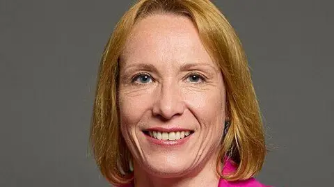 UK Parliament A posed shot in front of a grey background of a woman with blonde bobbed hair smiling. She has blue eyes and is wearing a pink jacket, which can just be seen.