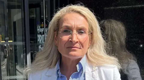 A woman with blonde hair and glasses, in a blue and white striped shirt and white jacket, stands by a black marble column with glass rotating doors in the background