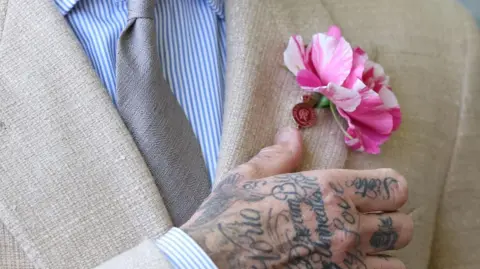 A pink and white striped flower in the lapel of a cream suit jacket with a tattooed hand reaching for it.
