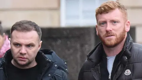 Tight head shot of two men walking outside a court building. One has brown hair, combed to the side, and the other has red hair and a beard.