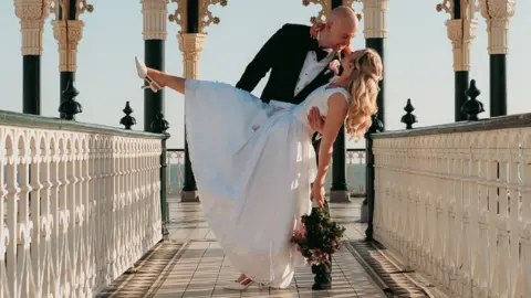 The image shows a groom holding his bride as she dips with one foot in the air and a bouquet in her hand. They are on what looks like a pier and the sky behind them is clear.