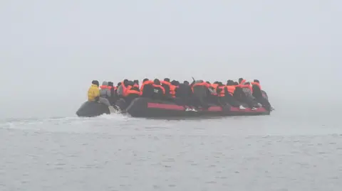 A small boat in the distance on the Channel. The boat is turned away from the camera, as migrants sit on either side of the boat, onwards to the UK.