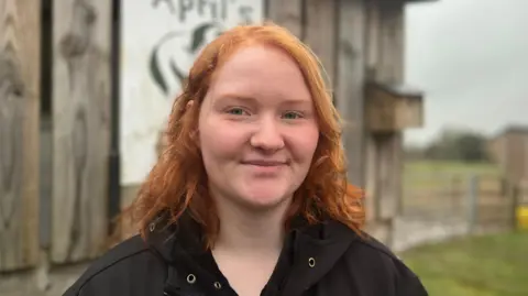 Emily has shoulder-length red hair and is wearing a black jacket with gold button holes. She is looking at the camera and behind her is a small wooden shelter, with a wooden birdhouse and fences and fields visible in the background. 