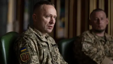 A man sits wearing a military uniform while another uniformed man looks on in the background