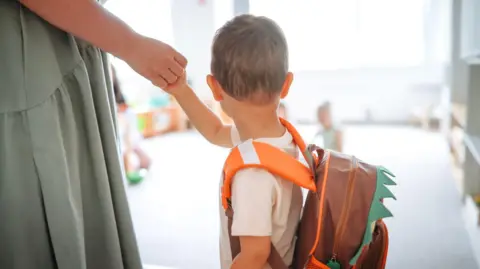 A young boy with his back to the camera holding a woman's hand in a nursery setting.  The boy has short, blonde hair and is wearing a white t-shirt and a brown backpack with orange straps. 