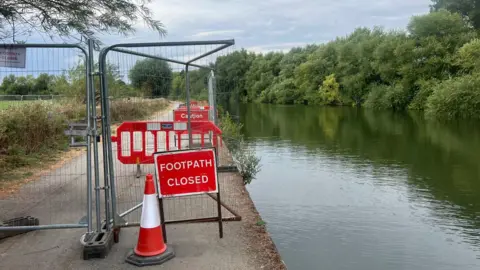 A footpath closed sign, wire fencing and a traffic cone mark the start of a path closure beside a river.