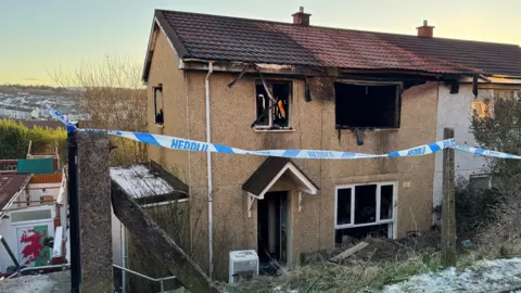 Burnt out end-of-terrace house with blackened windows and front door surrounded by police tape. The house is situated a little below street level with steps down. There is snow on the grassy verge above