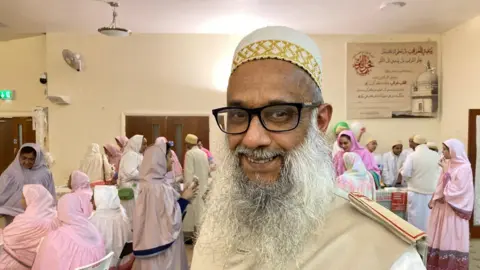 A man with a silver beard and wearing a white and gold embroidered Topi hat.