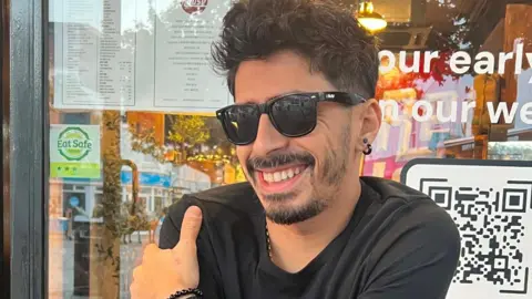 Mariano Ferreira Javier is wearing black sunglasses sits at an outdoor café table. His arm rests on the table near a stack of red-backed playing cards and a smartphone. Behind him, a window displays menus, signage, and a visible QR code.