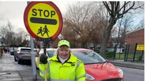 Margie is holding a stop sign and is dressed in full high visibility yellow school crossing patrol uniform. She stands on a pavement near a row of parked cars outside a school gates.