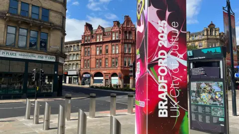 A poster that says Bradford 2025 UK City of Culture on a pillar with a fish and chip shop and a historic building in the background. 