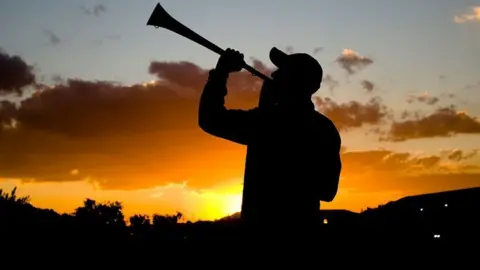 A silhouette of a football fan blowing a vuvuzela horn against the sunset