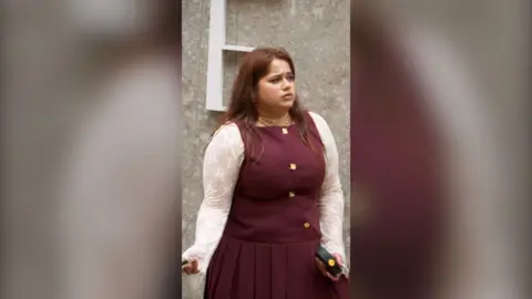 A woman in a burgundy dress with lace sleeves stands looking confused